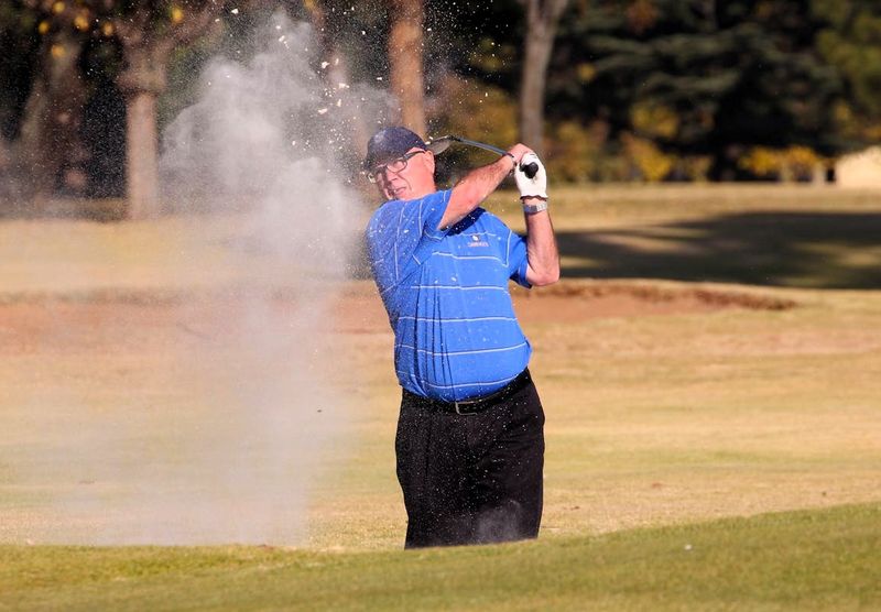 an older man wearing a blue golf shirt taking a golf shot from a bunker