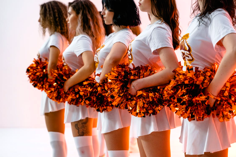 a squad of cheerleaders in white uniforms holding orange pom poms