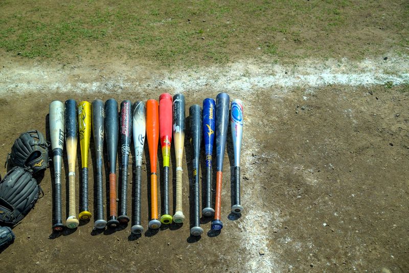 multiple sizes of baseball bats lying on a dirt baseball diamond sideline