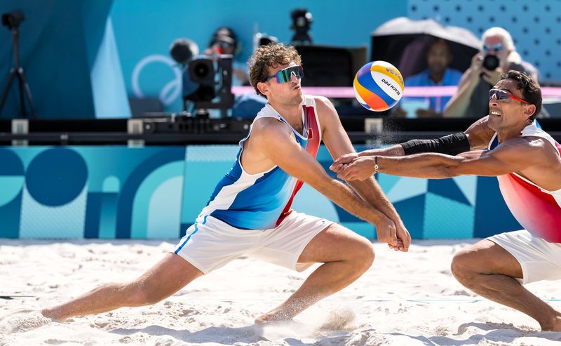 two male volleyball players both reaching for a volleyball on sand