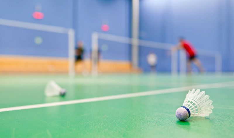 two shuttlecocks on an aqua color tennis court surface with badminton players in the backround