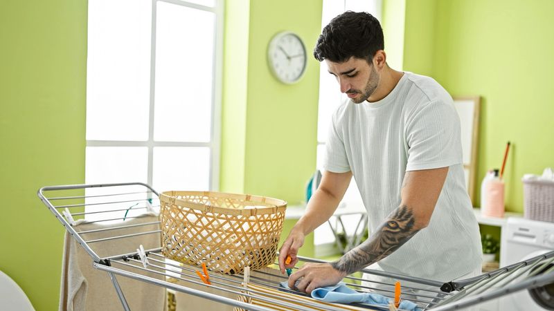 an adult male placing pegs on a softball jersey on a clothing drying rack in a laundry room
