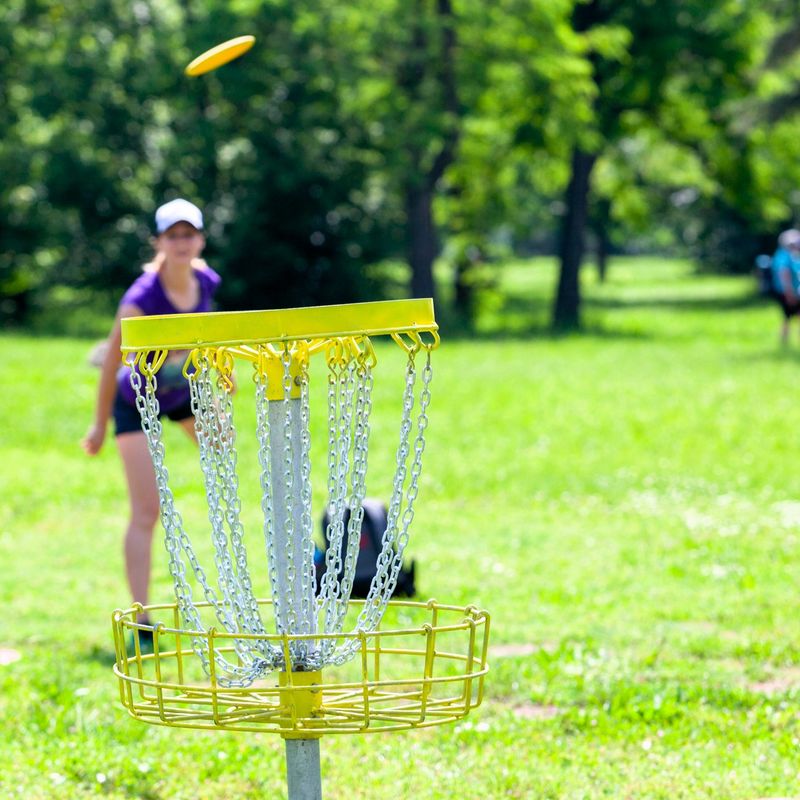an adult female disc golf player throwing a disc at a yellow disc golf bucket